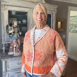 May include: A quilted jacket in shades of peach and orange featuring a floral pattern, a white tie-dye trim along the collar and sleeves, and a solid orange band at the hem. The jacket is worn over a white shirt and blue denim jeans.