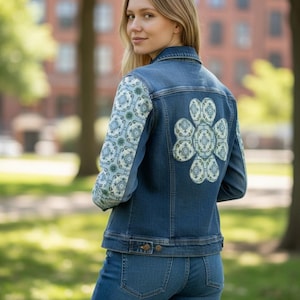 May include: A denim jacket and jeans ensemble. The jacket features patterned sleeves and a large floral design on the back. The sleeves and back design have a light blue and white pattern. The model is outdoors.