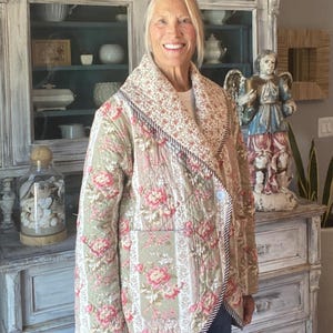 Peut inclure: Une veste matelassée avec un motif floral dans des tons vert sauge, rose et crème. Le vêtement présente un large col châle, des manches longues et une fermeture à un seul bouton. Les bords sont finis avec un liseré rayé marron et blanc.