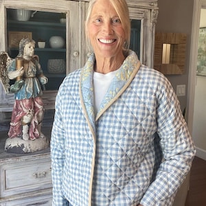 May include: A quilted jacket featuring a light blue and white gingham pattern. The jacket has a shawl collar lined with a light blue fabric displaying a yellow and white floral print, finished with a thin beige trim along the edges.