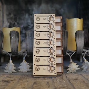 May include: A wooden daily pill organizer with seven compartments, each labeled with a day of the week: Sun, Mon, Tue, Wed, Thu, Fri, Sat. Each compartment has a sun and moon icon.