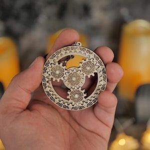 Op de afbeelding: Een houten sleutelhanger met drie in elkaar grijpende tandwielen, elk met een gedetailleerd ontwerp. De tandwielen zijn omgeven door een cirkelvormig frame met het woord "Allah" dat meerdere keren wordt herhaald.