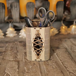 Puede incluir: Un porta lápices de madera con un diseño de encaje decorativo. El porta lápices está hecho de madera clara y tiene un acabado natural. Está lleno de bolígrafos y tijeras.