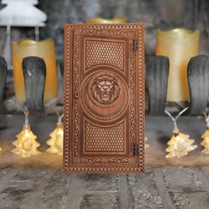 Pu&ograve; includere: Una scatola di legno con un design intricato che presenta una testa di leone al centro. La scatola ha un coperchio incernierato e un chiavistello.