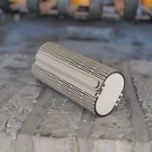 Op de afbeelding: Een cilindrische houten doos met een lasergesneden ontwerp. De doos is gemaakt van licht hout en heeft een natuurlijke afwerking. De doos is gesloten en ligt op een grijs bakstenen oppervlak.