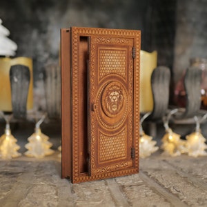 Pu&ograve; includere: Una scatola di legno con un design a testa di leone sul davanti. La scatola &egrave; aperta e l'interno &egrave; visibile. La scatola &egrave; in legno marrone chiaro e ha una macchia marrone scuro.