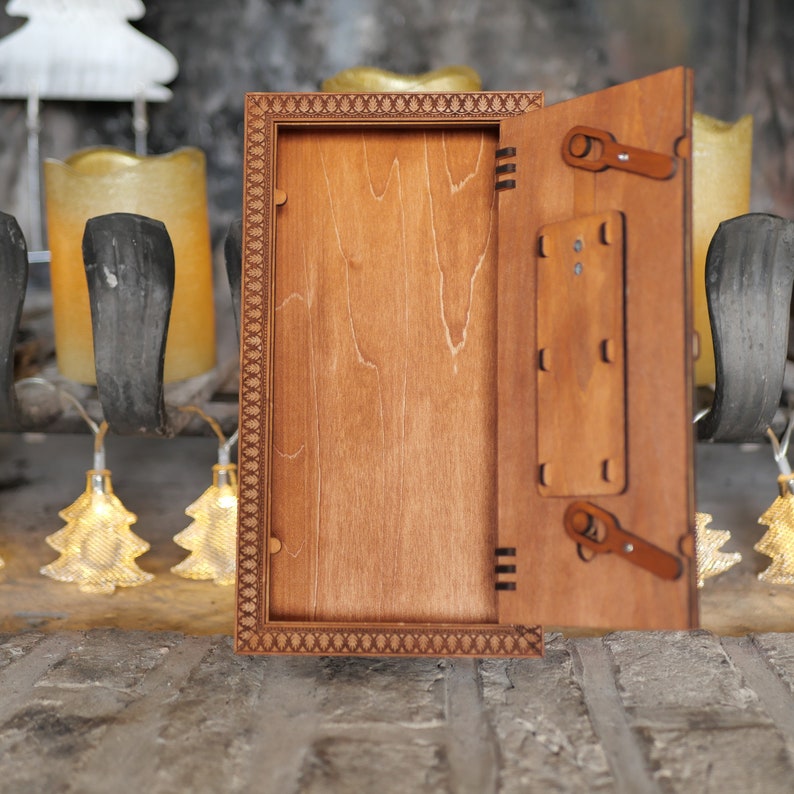 Pu&ograve; includere: Una scatola di legno con un coperchio incernierato che si apre per rivelare uno spazio vuoto all'interno. La scatola ha un bordo decorativo ed &egrave; realizzata in legno marrone chiaro.