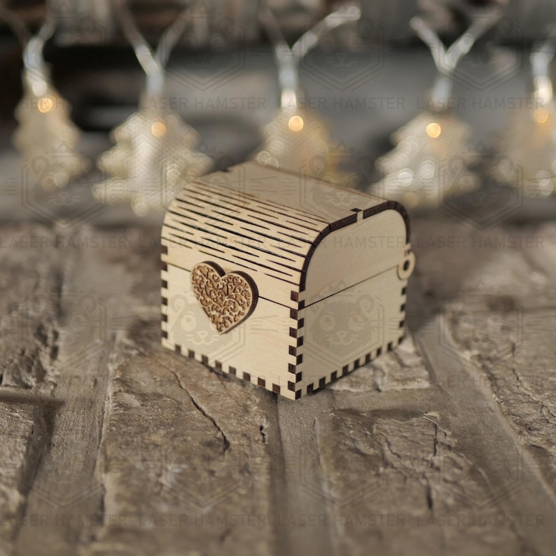 May include: A small, light-colored wooden chest with a heart-shaped embellishment. The chest has a curved top and is decorated with a laser-cut design. The heart is intricately carved with a floral pattern. The chest is sitting on a brick surface.