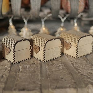 May include: Three small, wooden treasure chests with arched lids and heart-shaped accents. The boxes are light brown with a decorative pattern on the lid and a heart-shaped embellishment on the front. The chests are displayed on a textured surface.