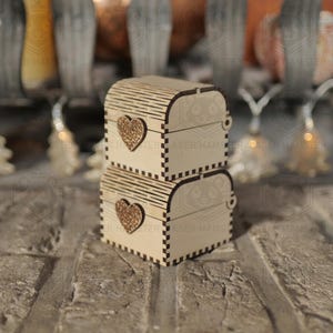 May include: Two small, wooden treasure chests stacked on top of each other. Each chest features a heart-shaped cutout on the front and a curved, slatted lid. The chests are made of light-colored wood and are set against a brick background.