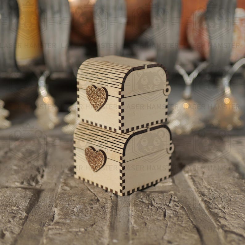 May include: Two small, wooden treasure chests stacked on top of each other. Each chest features a heart-shaped cutout on the front and a curved, slatted lid. The chests are made of light-colored wood and are set against a brick background.