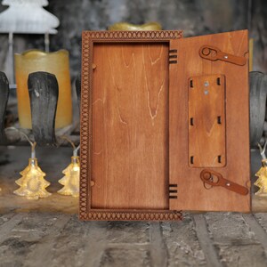 Pu&ograve; includere: Una scatola di legno con un coperchio incernierato, il coperchio &egrave; aperto rivelando l'interno della scatola. La scatola &egrave; in legno marrone chiaro con una macchia marrone scuro. La scatola ha un bordo decorativo attorno al bordo.