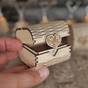 Puede incluir: Un pequeño cofre de madera abierto con un adorno en forma de corazón. El cofre está hecho de madera de color claro y presenta intrincados detalles cortados con láser. El corazón tiene un diseño floral. El cofre está abierto por una mano.