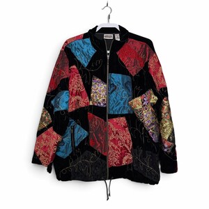 May include: A black zip-up jacket featuring a patchwork design of red, blue, and gold patterned fabric panels. The jacket includes a high collar, long sleeves, two front pockets with zippers, and a drawstring hem with black cord toggles.