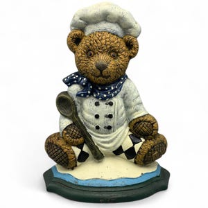 May include: A brown teddy bear figurine dressed as a chef, wearing a white hat, a white double-breasted jacket with black buttons, and a blue polka-dot neckerchief. The bear holds a wooden spoon and sits on a base with black and white checkered accents.