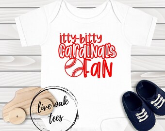 Könnte beinhalten: Weißer Baby-Strampler mit rotem Text "itty-bitty Cardinals Fan" und einer roten Baseball-Grafik. Der Strampler liegt auf einem Holz-Hintergrund mit einem Spielzeugauto und einem Paar Babyschuhen.