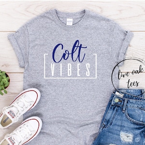 May include: Gray t-shirt with the text "Colt Vibes" in blue lettering inside a white box. The shirt is laid flat on a wood surface with a pair of white sneakers and denim shorts.