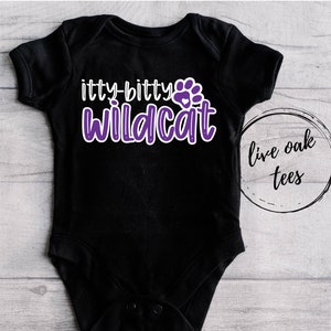 Op de afbeelding: Zwarte baby romper met paarse tekst die "itty-bitty wildcat" luidt, met een paarse voetafdruk met een wit hart in het midden. De romper ligt op een houten achtergrond met een witte cirkel waarop in zwarte tekst "live oak tees" staat.