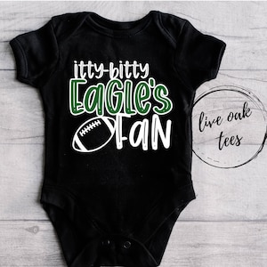 May include: Black baby onesie with white text that reads "itty-bitty Eagle's Fan" and a white football graphic. The text is in a green font. The onesie has a white circle on the right side with the text "live oak tees" in black.