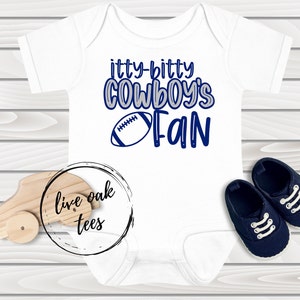 Peut inclure: Body bébé blanc avec l'inscription "itty-bitty COWBOY'S FAN" en bleu et gris. Un ballon de football américain bleu est en dessous. Des chaussures de bébé bleu marine et une voiture en bois sont à côté.
