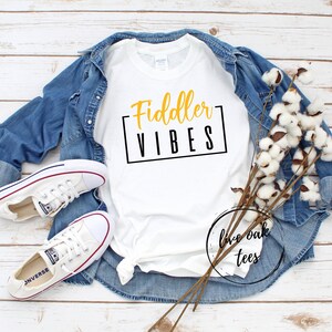 Może przedstawiać: Biała koszulka z napisem "Fiddler Vibes" w czarnych i złotych literach. Koszulka leży na drewnianej powierzchni z dżinsową kurtką i bawełnianymi łodygami.
