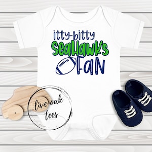 Op de afbeelding: Een witte baby romper met groene en blauwe tekst die "itty-bitty Seahawks Fan" luidt en een blauwe en witte voetbalafbeelding. De romper ligt op een houten achtergrond met een speelgoedauto en een cirkel met de tekst "live oak tees".