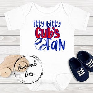 May include: White baby onesie with blue and red text that reads "itty-bitty Cubs Fan" and a baseball graphic. The onesie is on a wood background with a toy car and a logo that reads "live oak tees".