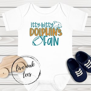 May include: White baby onesie with teal and orange text that reads "itty-bitty dolphin's fan". A teal dolphin graphic is next to the text. A football graphic is below the text. The onesie is on a wooden surface.