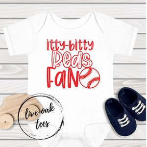 Puede incluir: Mono blanco para bebé con texto rojo que dice "itty-bitty Reds Fan" y un gráfico de béisbol rojo.