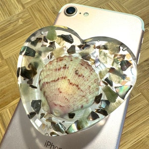 May include: A heart-shaped phone grip featuring a seashell encased in clear resin. The grip is adorned with iridescent and dark green fragments. The phone is a rose gold color.