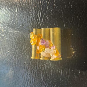 May include: Three brass bullet casings are adorned with colorful, translucent resin and small, irregularly shaped gemstones in shades of yellow, purple, and white. The arrangement suggests a unique, handcrafted decorative item.