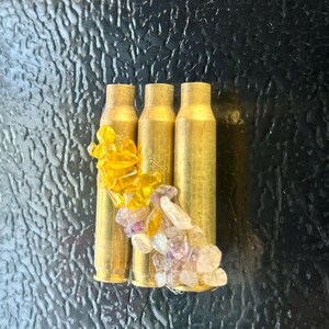 May include: Three brass bullet casings adorned with yellow and purple gemstones. The casings are arranged vertically, with the gemstones clustered around them. The background is a textured, dark surface.