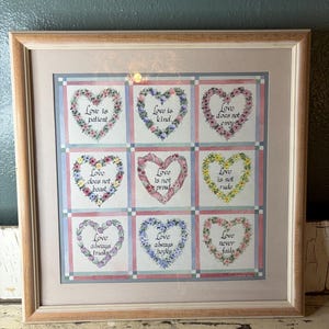 May include: A framed wall art piece featuring a grid of nine squares, each containing a heart-shaped floral wreath surrounding text. The phrases include Love is patient, Love is kind, Love does not envy, Love does not boast, Love is not proud, Love is not rude, Love always trusts, Love always hopes, and Love never fails.