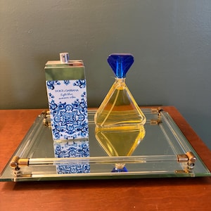 May include: A rectangular Dolce & Gabbana Light Blue Summer Vibes perfume bottle with a blue and white floral pattern and a triangular perfume bottle with a blue diamond-shaped cap and yellow liquid, both resting on a square mirrored tray.