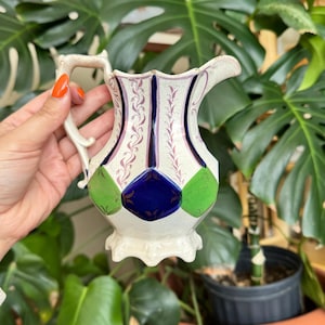 Pode incluir: Um jarro de cerâmica branca com um padrão geométrico azul e verde. O jarro tem uma alça e uma base festoneada.