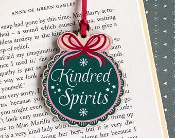 May include: A wooden Christmas ornament with a green center and the words "Kindred Spirits" in white script. The ornament has a pink and white scalloped border and a red ribbon for hanging.