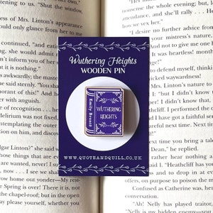 Puede incluir: Un pin de madera con un libro morado con el t&iacute;tulo "Wuthering Heights" y el autor "Emily Bront&euml;" impreso en la portada. El pin est&aacute; sobre un fondo morado con el texto "Wuthering Heights WOODEN PIN" y el sitio web "www.QUOTEANDQUILL.CO.UK".