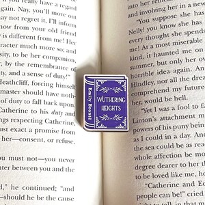 Puede incluir: Un pin de madera de color morado con forma de libro con el t&iacute;tulo "Wuthering Heights" y el autor "Emily Bront&euml;" impreso en &eacute;l. El pin est&aacute; sobre un libro abierto.