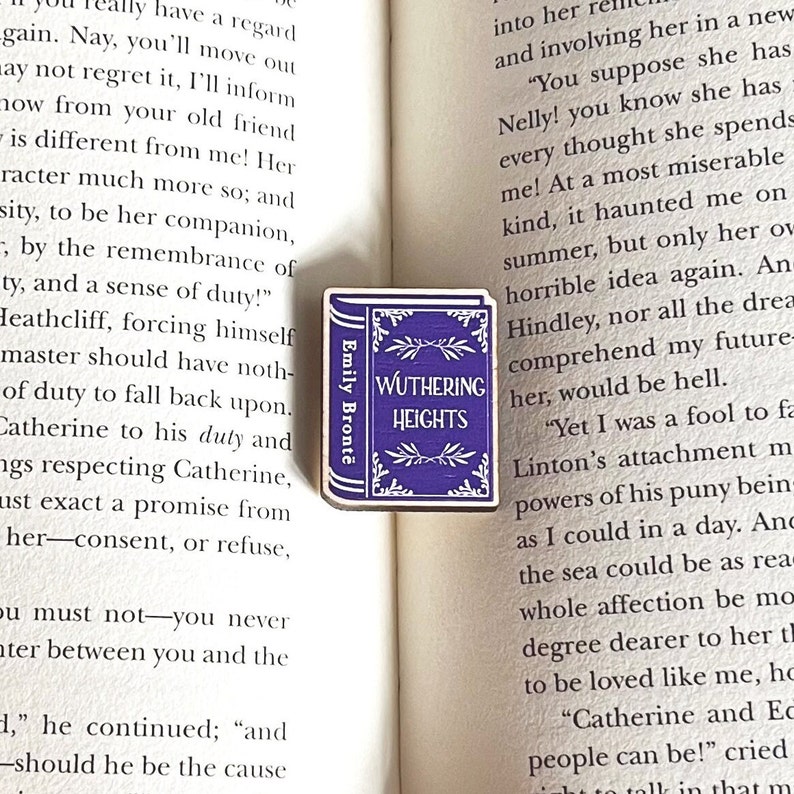 Puede incluir: Un pin de madera de color morado con forma de libro con el t&iacute;tulo "Wuthering Heights" y el autor "Emily Bront&euml;" impreso en &eacute;l. El pin est&aacute; sobre un libro abierto.