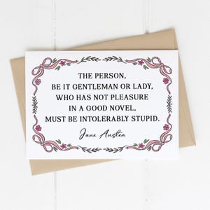 May include: A white card featuring a quote by Jane Austen, surrounded by pink ribbon and floral decorations. The quote states: "THE PERSON, BE IT GENTLEMAN OR LADY, WHO HAS NOT PLEASURE IN A GOOD NOVEL, MUST BE INTOLERABLY STUPID." The card is on a beige envelope.