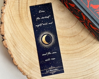 May include: A navy blue bookmark with gold text and a crescent moon design. The bookmark reads "Even the darkest night will end and the sun will rise" and includes the text "Les Misérables".