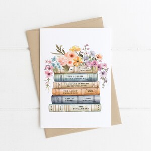 May include: A greeting card featuring a watercolor illustration of a stack of books with floral accents. The books are in various colors, with titles like "The Custom of the Country" and "The Age of Innocence". A brown envelope is behind the card.