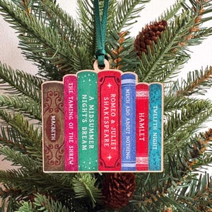 May include: A flat wooden ornament shaped like a row of seven books with a dark green ribbon loop. The spines are labelled with titles including Macbeth, The Taming of the Shrew, A Midsummer Night's Dream, Romeo & Juliet, Much Ado About Nothing, Hamlet, and Twelfth Night.