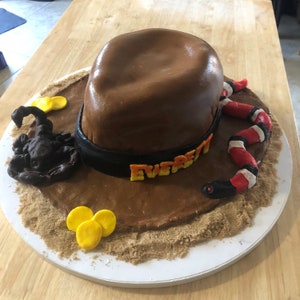 Pode incluir: Um bolo castanho e preto decorado para parecer um chapéu de cowboy. O chapéu tem uma faixa preta com o nome "EVERETT" escrito em letras laranja e amarelas. O bolo é rodeado por açúcar mascavo para parecer areia. Há um escorpião preto e uma cobra vermelha e branca no bolo.