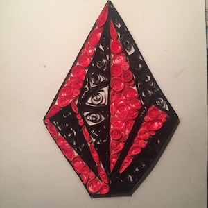 May include: A red and black paper quilling art piece in the shape of a diamond with a swirling eye design in the center.