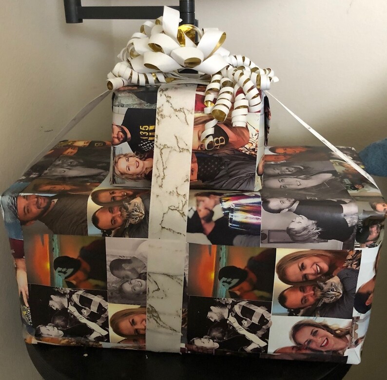 Op de afbeelding: Een cadeaudoos verpakt in wit papier met een marmerpatroon en een wit lint. De doos is versierd met foto's van mensen en heeft een gouden strik bovenop.