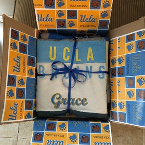 Op de afbeelding: Een cadeaudoos ingepakt in geel en blauw gepatroneerd papier met een blauw lint. Het papier heeft het UCLA Bruins-logo en de woorden "UCLA Bruins". De doos bevat een wit deken met het woord "Grace" in gouden letters.