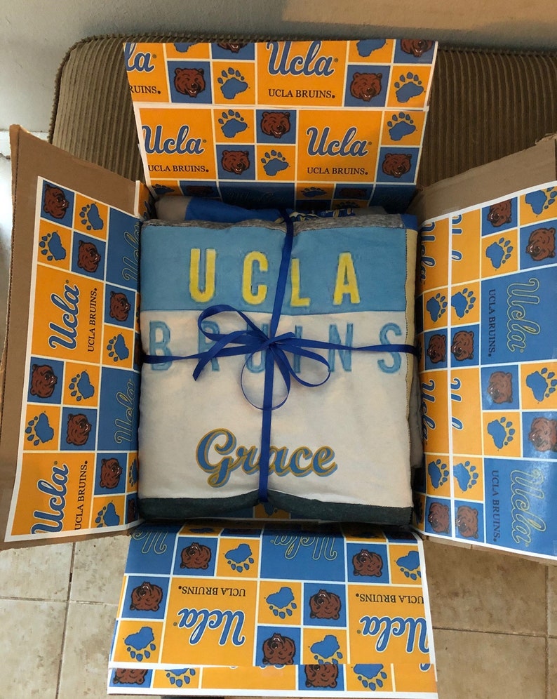 Op de afbeelding: Een cadeaudoos ingepakt in geel en blauw gepatroneerd papier met een blauw lint. Het papier heeft het UCLA Bruins-logo en de woorden "UCLA Bruins". De doos bevat een wit deken met het woord "Grace" in gouden letters.