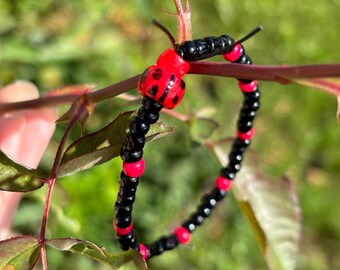 Könnte beinhalten: Ein Perlenarmband mit einem Marienkäfer-Design. Das Armband besteht aus schwarzen und roten Perlen, mit einem roten Marienkäfer-Anhänger in der Mitte. Das Armband wird auf einem Rosenzweig vor einem verschwommenen grünen Hintergrund präsentiert.