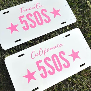 May include: Two rectangular white number plates with rounded corners and four mounting slots. Each plate features the pink text 5SOS flanked by two pink stars, with the word Toronto on one plate and California on the other in pink script.